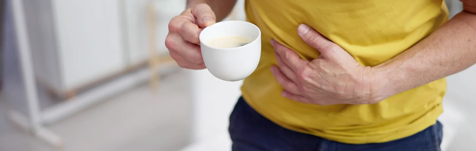 Męczy Cię zgaga po kawie? Odczuwasz pieczenie w żołądku? Sprawdź, dlaczego czasami po kawie boli brzuch. Mężczyzna w żółtej koszulce łapie się za brzuch po wypiciu filiżanki kawy.